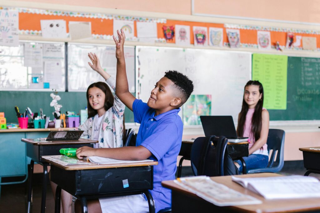 Children in a lively classroom raising hands during an interactive lesson.